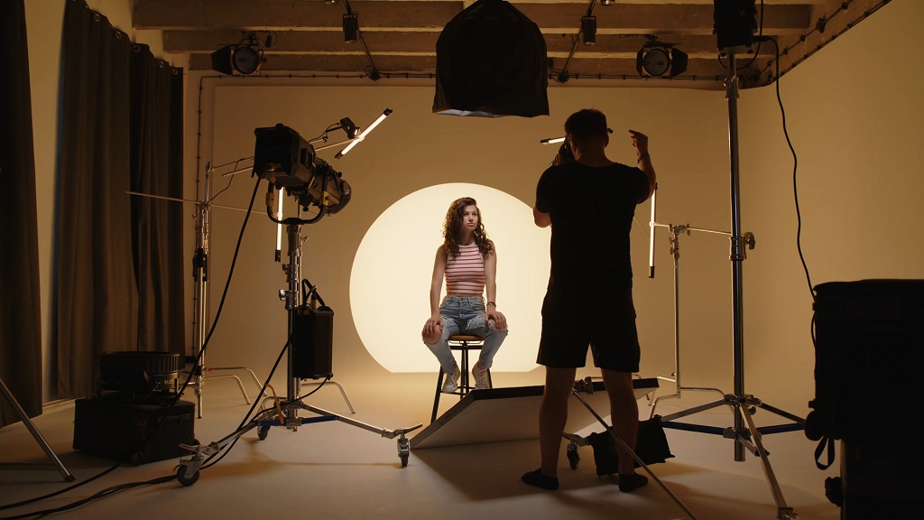 Model sedi na stolici u profesionalnom foto studiju dok fotograf podešava rasvetu ispred nje, sa kružnim svetlosnim akcentom u pozadini