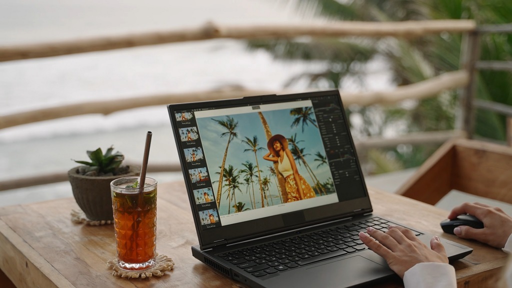 Osoba uređuje fotografije na laptopu dok sedi za drvenim stolom pored mora